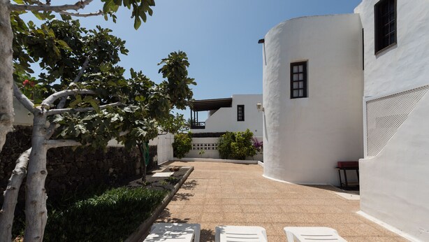 Un bâtiment blanc à la façade incurvée, doté d’un mur en pierre et d’un petit jardin avec un arbre et des plantes. On aperçoit deux fauteuils de jardin blancs au premier plan.