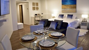 A modern hotel room with a glass dining table set for four, a sofa bed with multiple pillows, a painting on the wall, and a small desk with a chair. - Cozy Beachfront Condo With Jacuzzi On Balcony (Tambon Cha-am)
