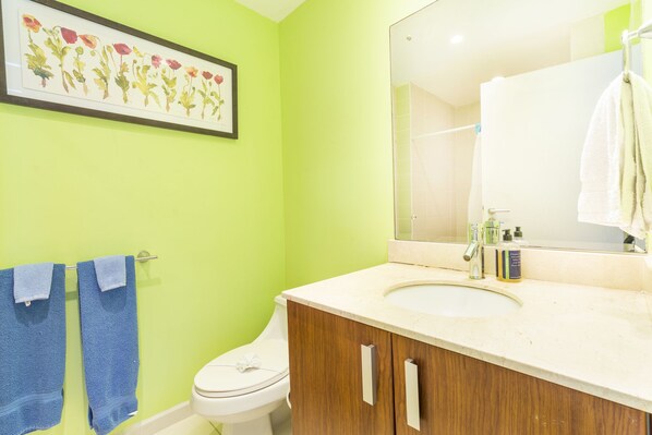 A bathroom with a green wall, a white toilet, a wooden cabinet with a sink, and a framed floral artwork above the toilet. - Vacation in a Beautiful 2Bed2Bath Condo,Jaco Beach (Jacó)