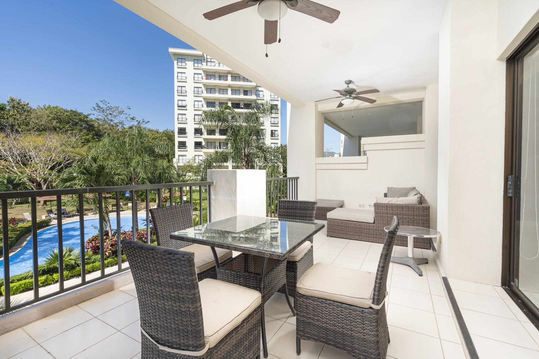 A balcony with wicker furniture, a glass table, and a view of a swimming pool and high-rise buildings.