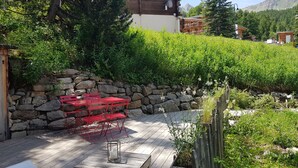 A wooden deck with red chairs and a table, surrounded by a stone wall and lush greenery. - Vars. Full-service center.  Private parking. Linens provided, beds made. (Vars)
