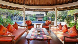 A covered outdoor seating area with wicker furniture and orange cushions, a table with a colorful table runner, and a view of a pool and greenery.