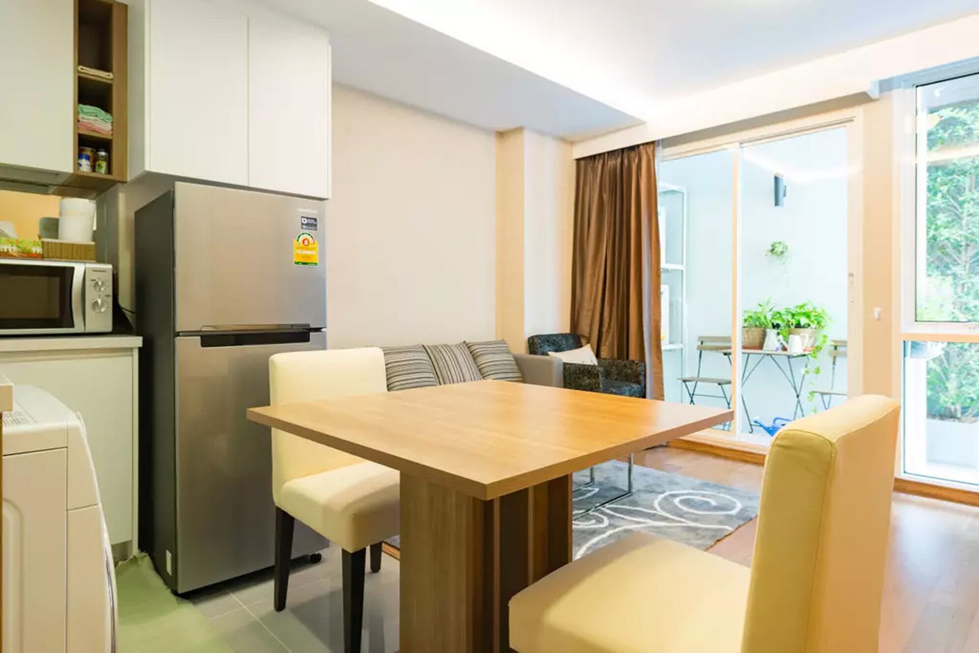 Eine kompakte Küche mit Kühlschrank, Mikrowelle und einem kleinen Schrank. Es gibt einen Essbereich mit einem Holztisch und Stühlen. Eine Glasschiebetür führt zu einem Balkon mit Topfpflanzen.