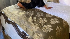 Une personne est assise sur un lit recouvert d’une couvre-lit à motifs. On aperçoit un lustre et une fenêtre avec des rideaux.