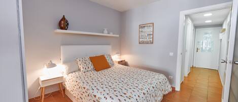 A bedroom with a bed, bedside tables, a nightstand, a shelf with decorative items, a wall-mounted sign, and a hallway leading to a door.