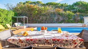 A dining table with a floral tablecloth, surrounded by wicker chairs, near a pool with a view of trees and a gazebo. - Villa Oásis, Piscina Privada, Ténis, Jacuzzi e Piscina Aquecida do Condomínio! (PortimÃ£o)