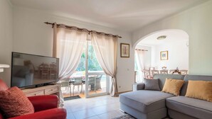 A living room with a TV, red and grey sofas, a dining area with chairs, and a large window with curtains. - Villa Oásis, Piscina Privada, Ténis, Jacuzzi e Piscina Aquecida do Condomínio! (PortimÃ£o)