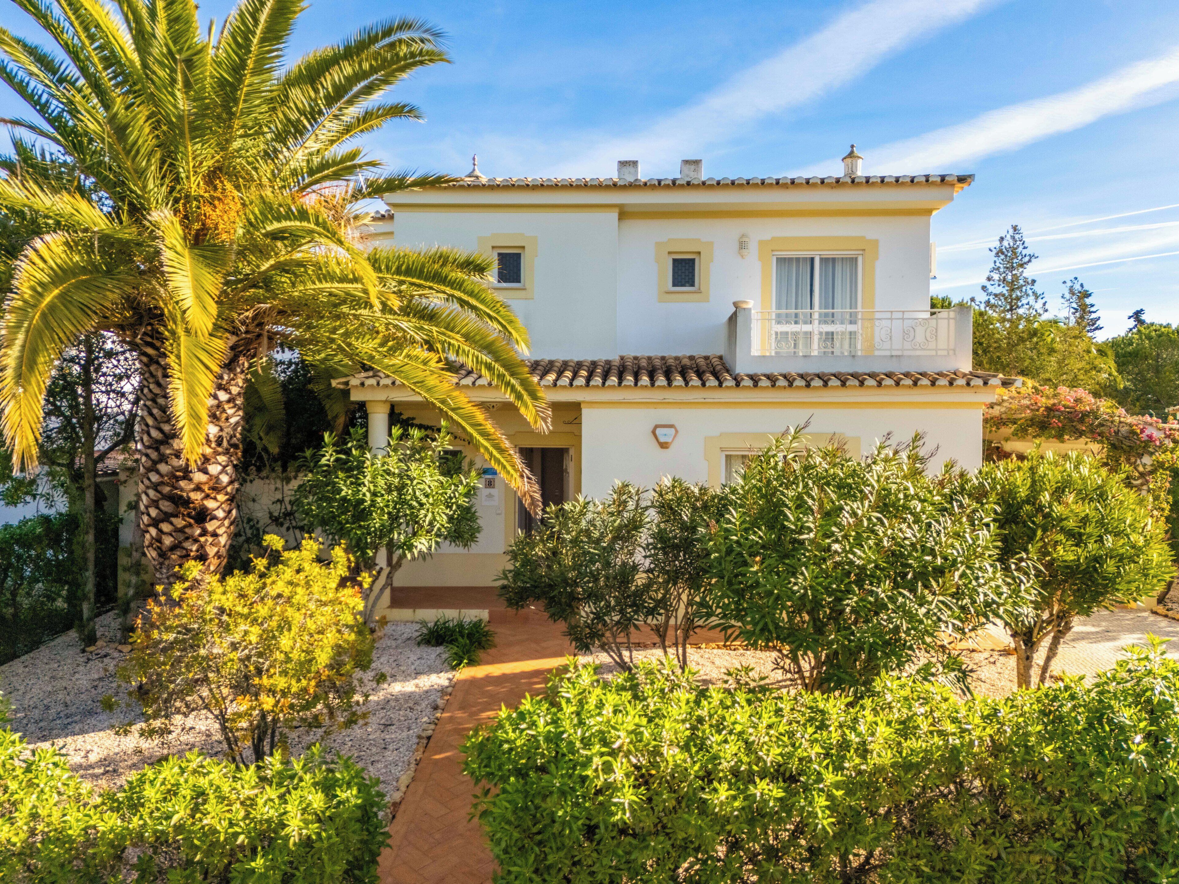 A two-story house with a balcony, surrounded by lush greenery and a palm tree.