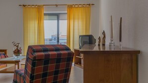 Una stanza con una poltrona a quadri, una scrivania in legno, una televisione e vista su un paesaggio attraverso una finestra con tende gialle.