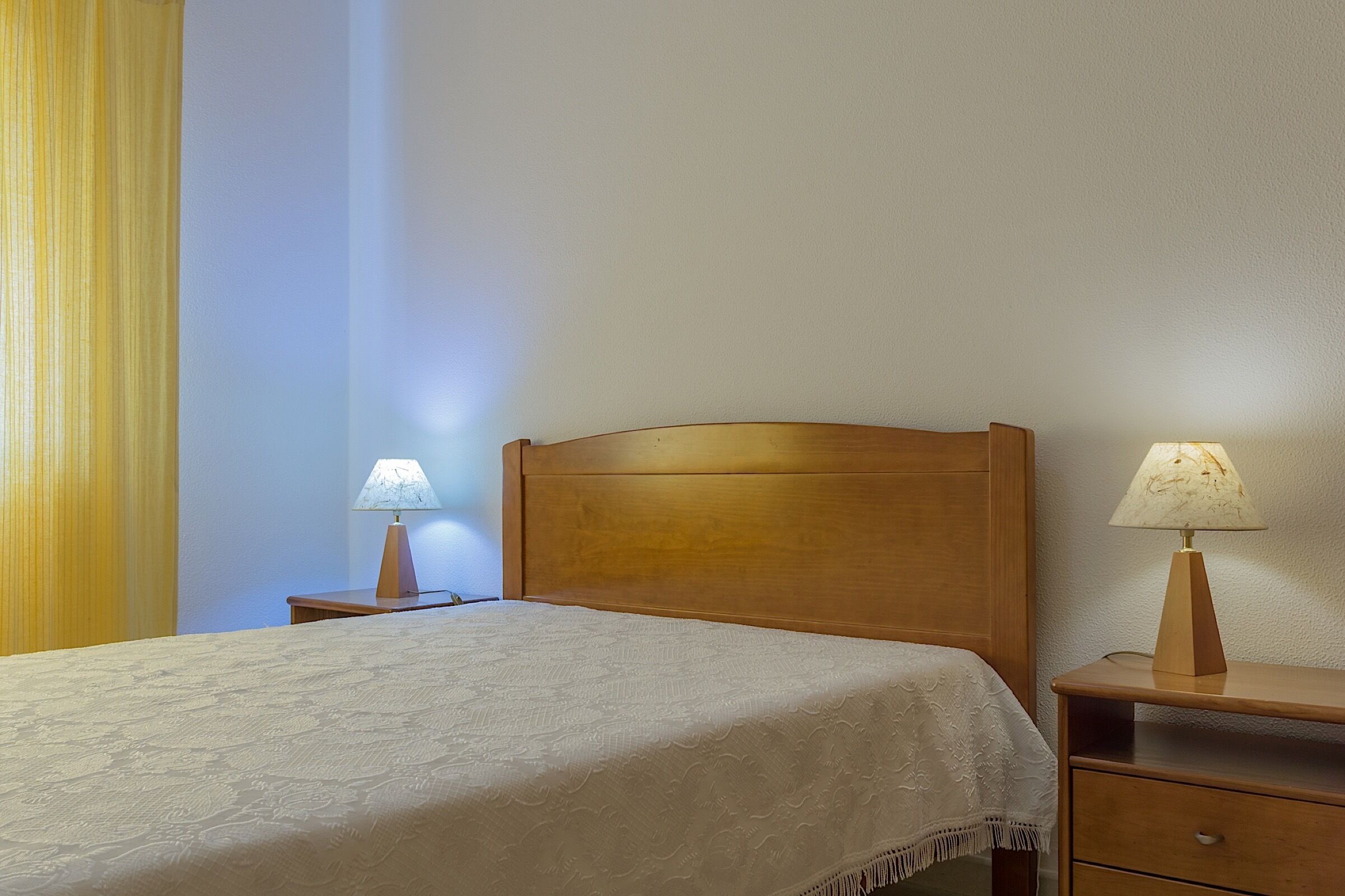 A neatly made bed with a wooden headboard, two bedside tables with lamps, and a plain wall.
