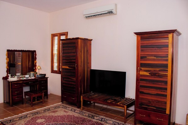 A living room with a TV, wooden furniture, a patterned rug, and an air conditioner. - BRIDAL SUITE - KING Room (Orchid Bay)