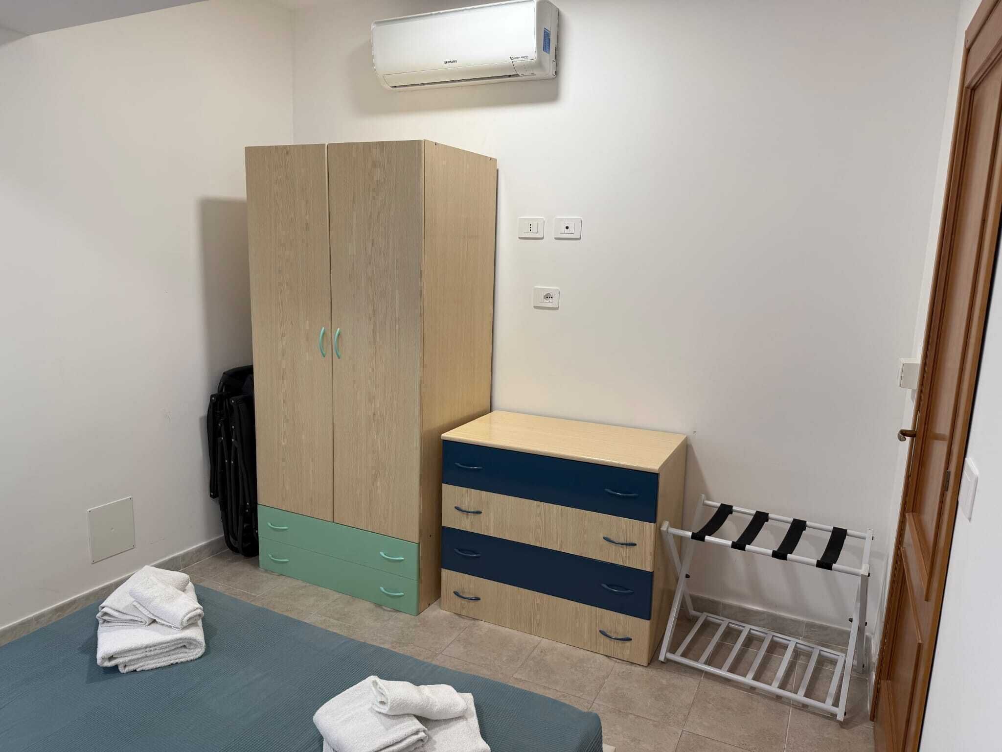 A bedroom with a wardrobe, a bed, a drying rack, and an air conditioning unit.