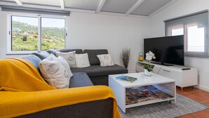 A modern living room with a grey sofa, a yellow blanket, a white coffee table with magazines, a flat-screen TV, and a view of greenery outside. - Apartment Casa Anny 2 (Madeira)