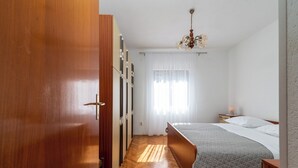 Une chambre avec une armoire en bois, un lit recouvert d’une courtepointe, une table de chevet avec une lampe et une fenêtre ornée de rideaux légers.
