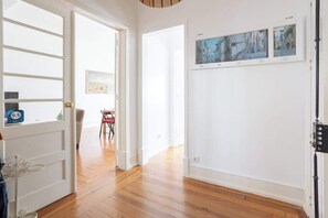 Un corridoio con pavimento in legno, pareti bianche e una porta con maniglia in metallo. Alle pareti si vedono quadri incorniciati e, sullo sfondo, una sedia.