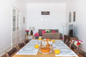 Zona pranzo con un tavolo lungo apparecchiato per un pasto, un letto con cuscini, una TV e un cartello appeso al muro.