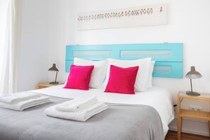 A neatly made bed with white and pink pillows, a grey blanket, and folded white towels on a white bedspread.
