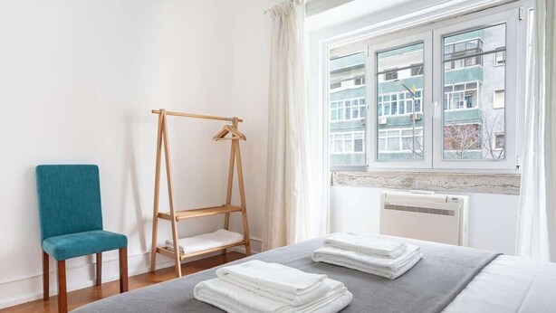 Hotellihuone, jossa on sänky, puinen pyyheteline, turkoosinsininen tuoli ja ikkuna, josta on näkymä rakennuksiin.