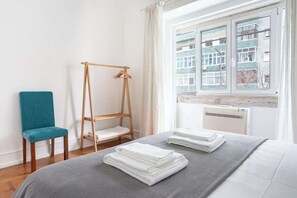 Une chambre d’hôtel avec un lit, un étendoir en bois, une chaise de couleur turquoise et une fenêtre donnant sur des immeubles.