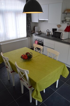 A compact kitchen with white cabinets, a microwave, a kettle, and a table set with a green tablecloth and chairs.