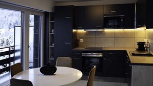 A modern kitchen with dark cabinetry, a round dining table, and a view of a snowy landscape through large windows. - PRETTY APARTMENT 4 to 6 people (Nendaz)
