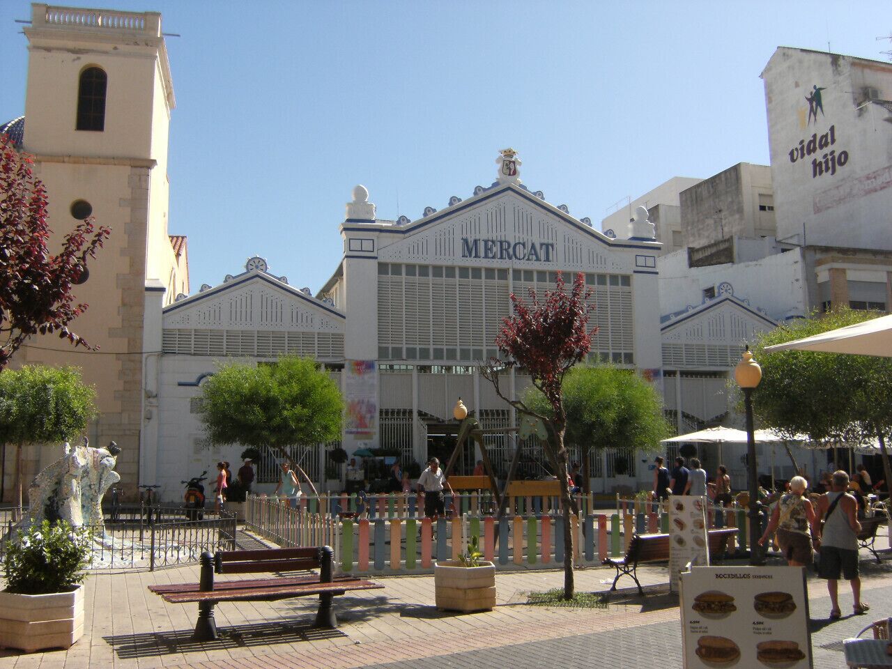 A Mercat building with a clock tower, surrounded by outdoor seating areas and potted plants.