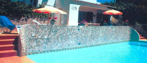 A swimming pool with a tiled edge, a stone wall, and a building with a chimney in the background.
