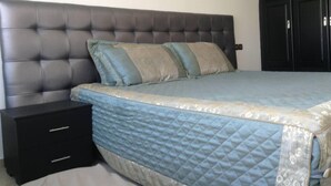 Ein Schlafzimmer mit einer gepolsterten Kopfstütze, einem Bett mit blauer Steppdecke, zwei Nachttischen und einem Schrank mit Spiegel.