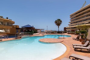 A swimming pool with sun loungers and a covered area, surrounded by buildings with balconies.