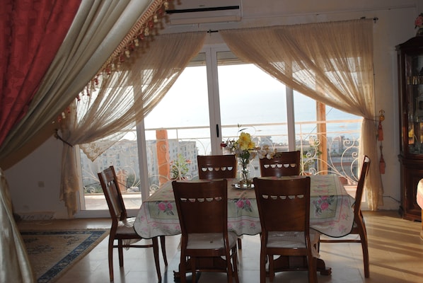 A dining area with a table set for four, surrounded by wooden chairs, a floral tablecloth, and a view of the city through large windows.