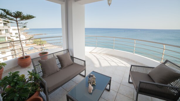 Een balkon met een bank en fauteuils, planten in potten en uitzicht op zee.