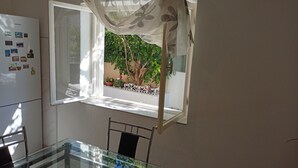 Uma área de refeições com mesa de vidro, uma cadeira, janela com cortina leve e uma geladeira com imãs.