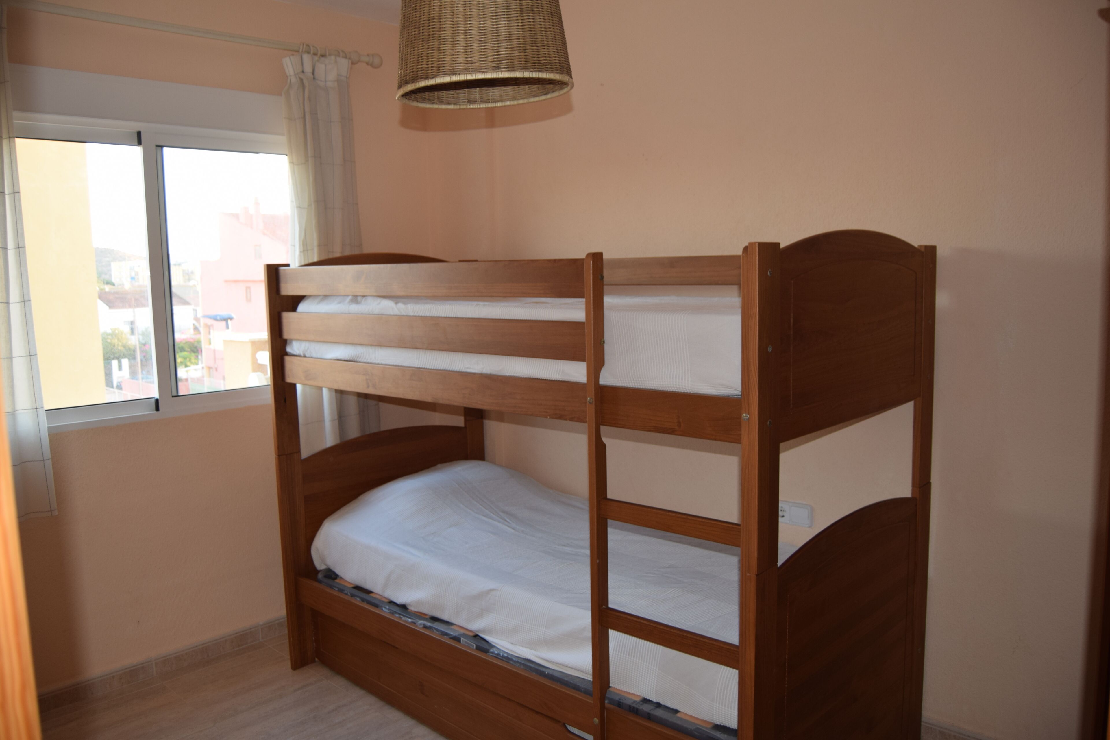 A bunk bed with a wooden frame and white bedding, a window with curtains, and a wicker lamp.