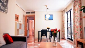 Ein Wohnzimmer mit einer Couch, einem Esstisch mit Stühlen, einer Vase mit Blumen und einem Fenster mit Vorhängen.