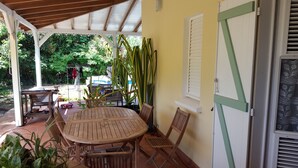 A covered patio area with a wooden table and chairs, surrounded by potted plants and a view of a pool.