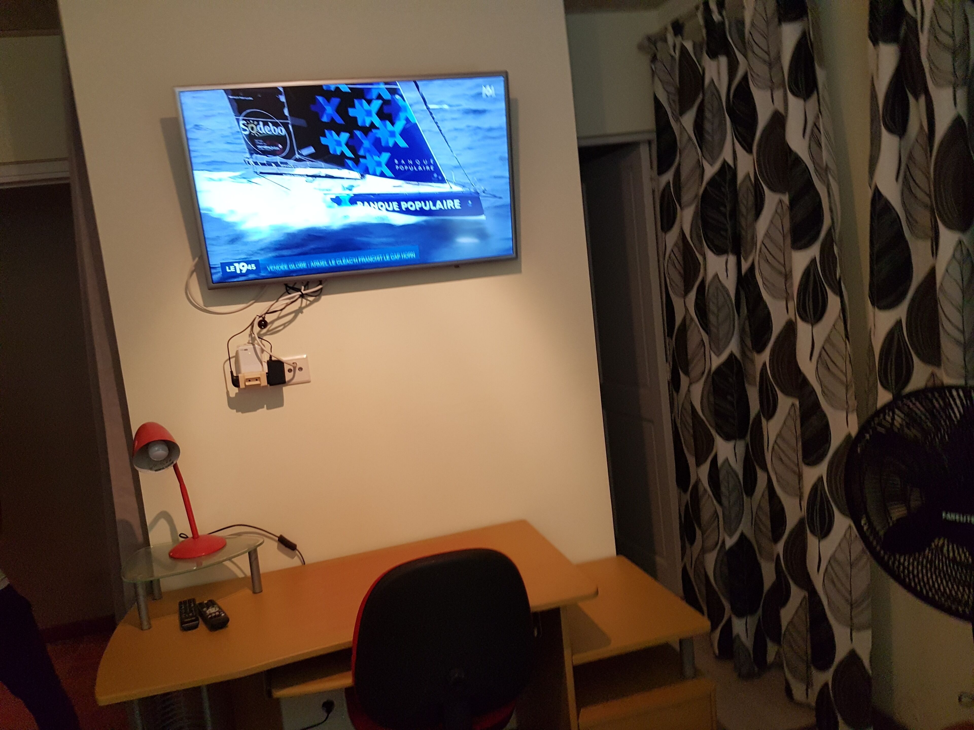A television mounted on a wall displaying a sports channel, a desk with a chair, a lamp, and a power outlet.
