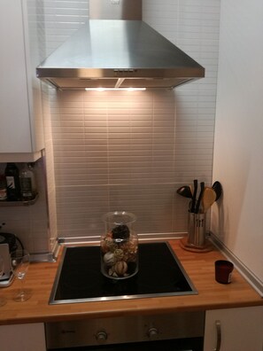Uma cozinha com coifa de aço inoxidável, um pote de vidro com objetos decorativos na bancada e um porta-utensílios com colheres e uma escova.