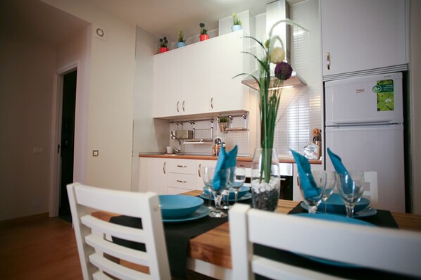 Uma área de refeições com mesa de madeira, cadeiras brancas e cozinha ao fundo.