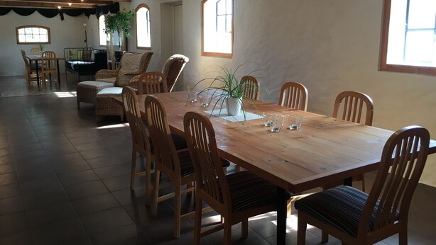 Un vaste espace dédié à la salle à manger, avec une grande table en bois et plusieurs chaises en osier. Des plantes en pot sont disposées sur la table, et la pièce est dotée de hauts plafonds avec des poutres apparentes.