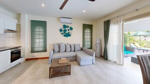 A modern living room with a grey sofa, a wooden coffee table, and a kitchen area with white cabinets and a stove. - Junior Pool Villa in Beach resort (Ko Samui)