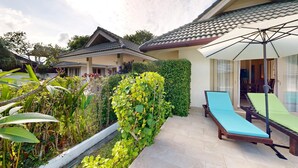 A villa with a tiled roof, a covered outdoor seating area with a lounge chair, and a lush garden with various plants. - Junior Pool Villa in Beach resort (Ko Samui)