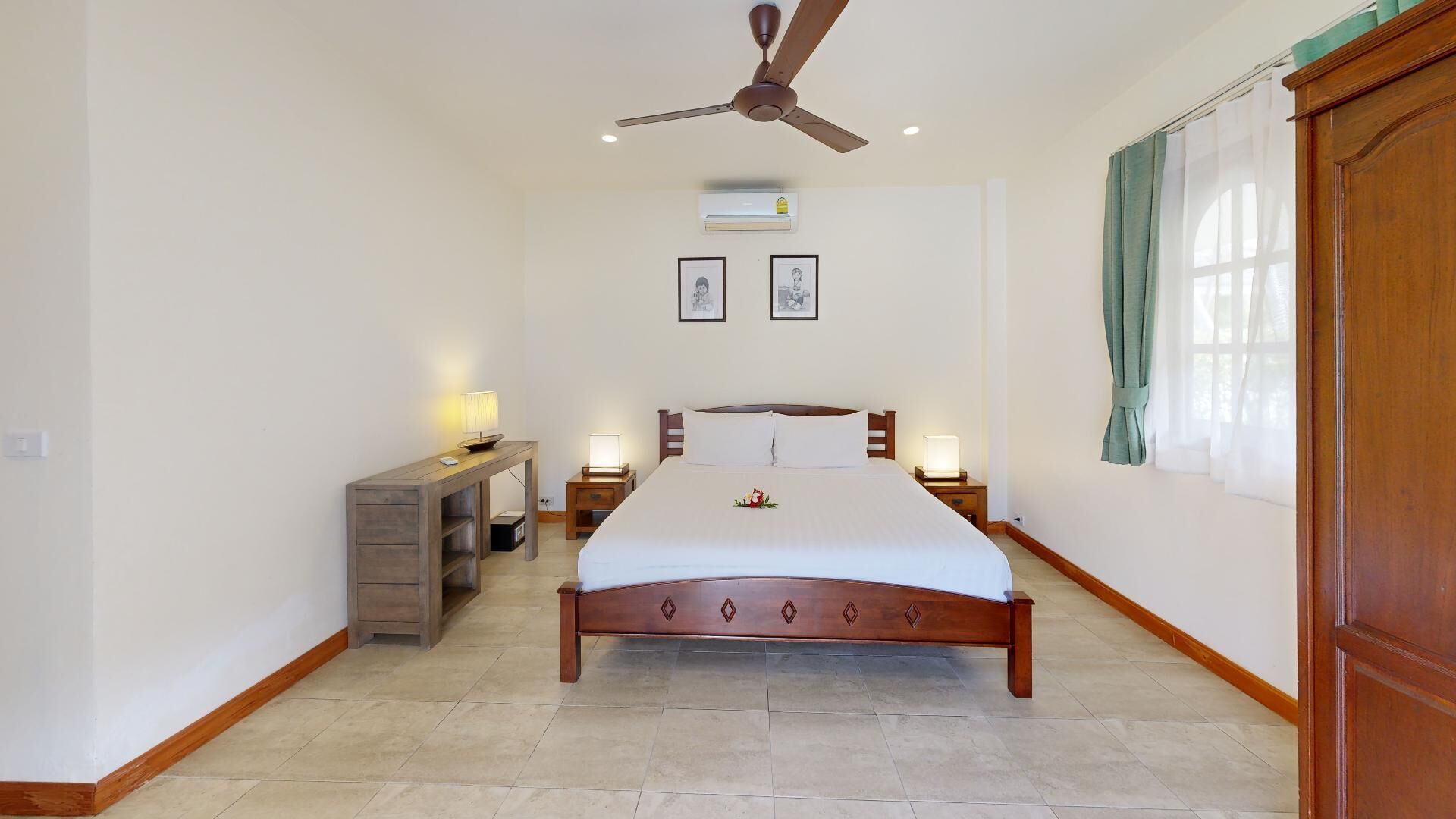 A bedroom with a wooden bed, a nightstand with a lamp, a desk with a lamp, a ceiling fan, and framed pictures on the wall.