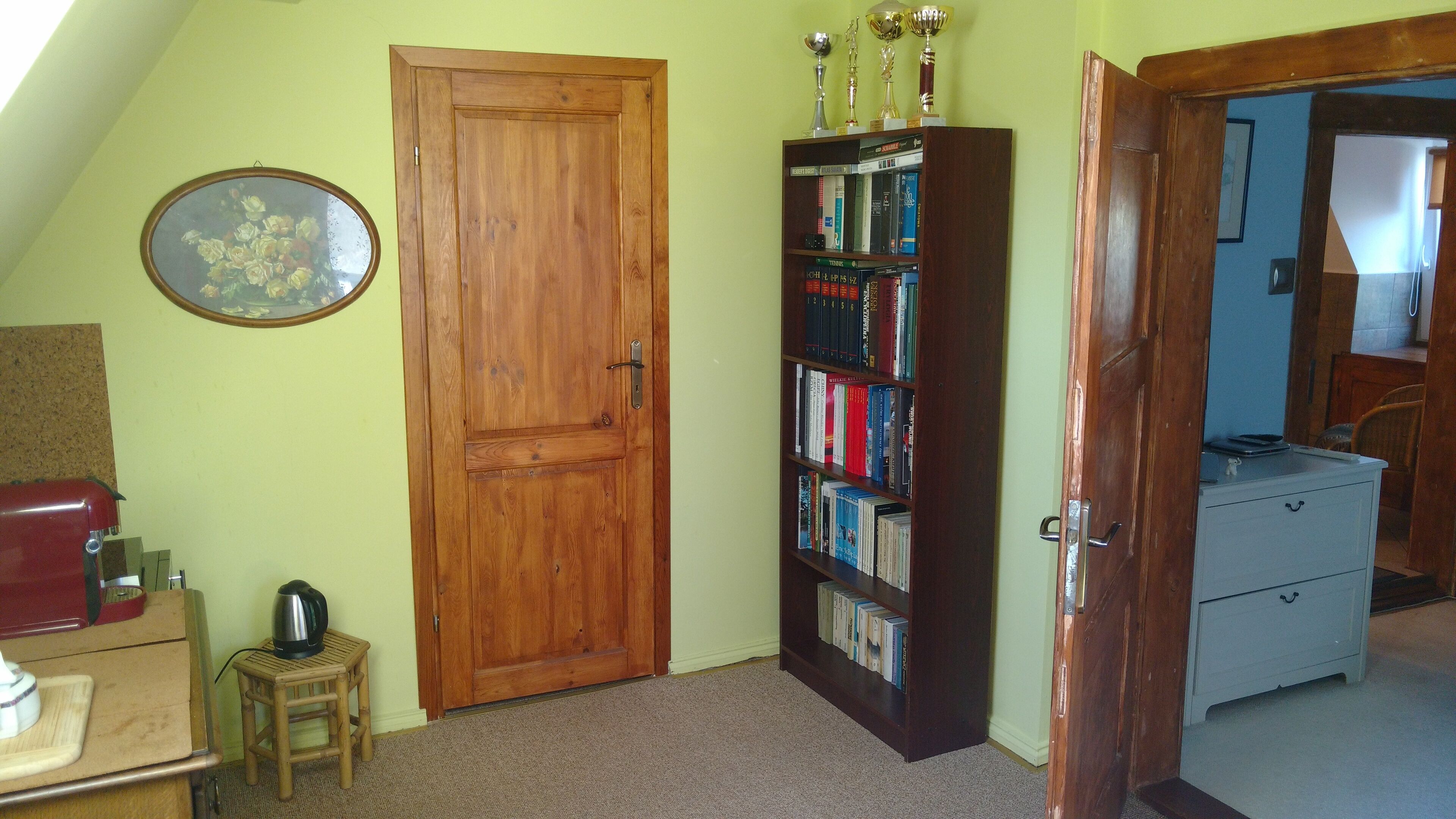 Um quarto com porta de madeira, estante, um banquinho pequeno e uma escrivaninha com chaleira.