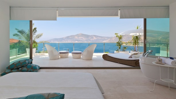 A bedroom with a large bed, a bathroom with a bathtub, and a balcony with seating and a view of the sea.