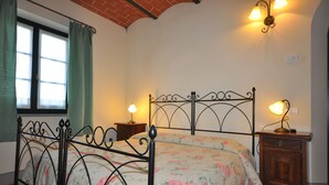 Ein Schlafzimmer mit Metallbettgestell, floralem Bettzeug, einem Nachttisch mit Lampe und einem Fenster mit Vorhängen.