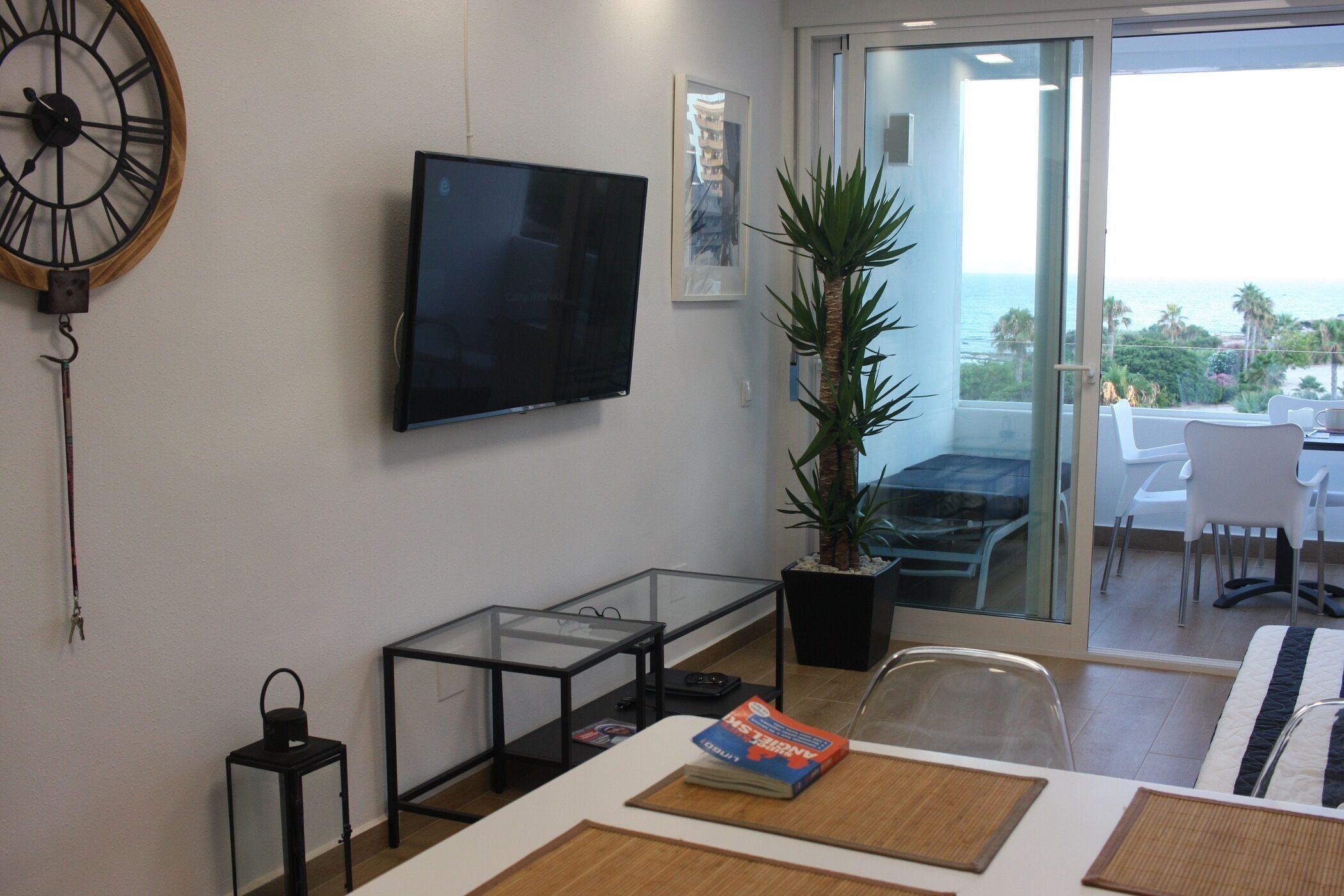 A modern living room with a flat-screen TV, a large wall clock, a glass coffee table, a potted plant, and a view of the ocean through a sliding glass door.