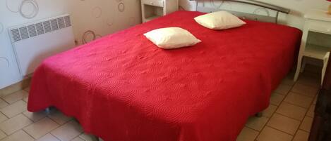 A bedroom with a red bedspread, two pillows, a bedside table with a lamp, and a radiator on the wall.