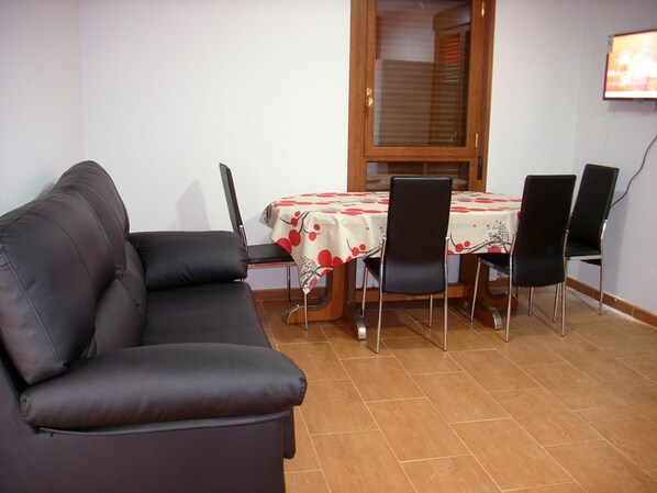 Un comedor con una mesa cubierta con mantel rojo y blanco, rodeada por cuatro sillas negras. Un sofá de cuero marrón se encuentra frente a la mesa.
