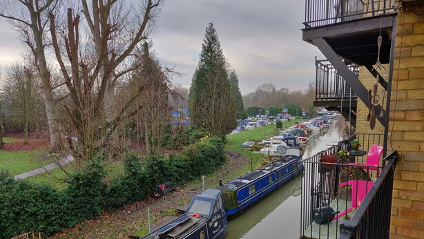 A narrow canal with a narrowboat docked alongside, a bench, and a building with a balcony. - Waterfront apartment in the Heart of St Neots (St Neots, Cambs)