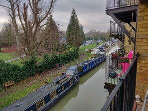 A narrow canal with a narrowboat docked alongside, a bench, and a building with a balcony. - Waterfront apartment in the Heart of St Neots (St Neots, Cambs)